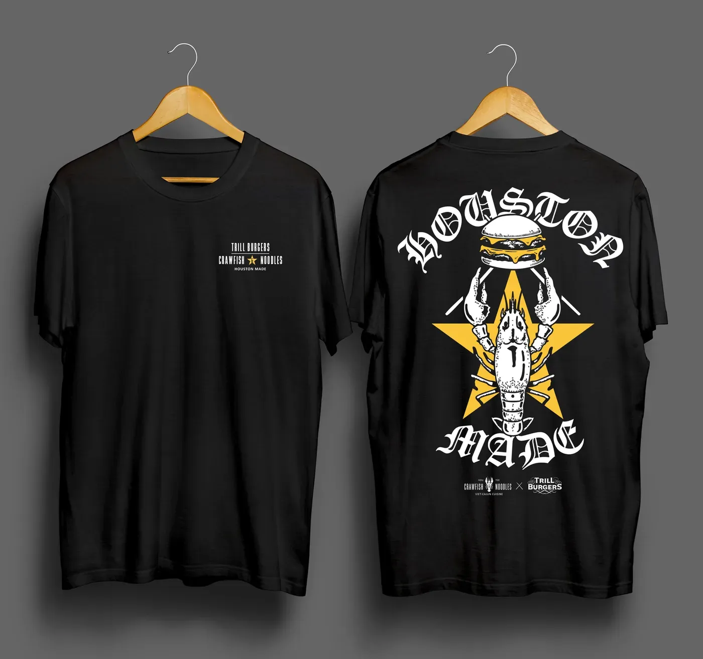 Front and back of a black t-shirt on wooden hangers against a grey backdrop. The front carries a small white chest lockup reading Trill Burgers above Crawfish and Noodles, with an ampersand set inside a yellow star, and Houston Made below. The back carries blackletter Houston arched above and Made curved below a white crawfish that raises a cheeseburger in its claws over a large yellow star, with four white bars radiating diagonally from behind it. Under the crest the Crawfish and Noodles logo and the Trill Burgers logo sit on either side of an X.