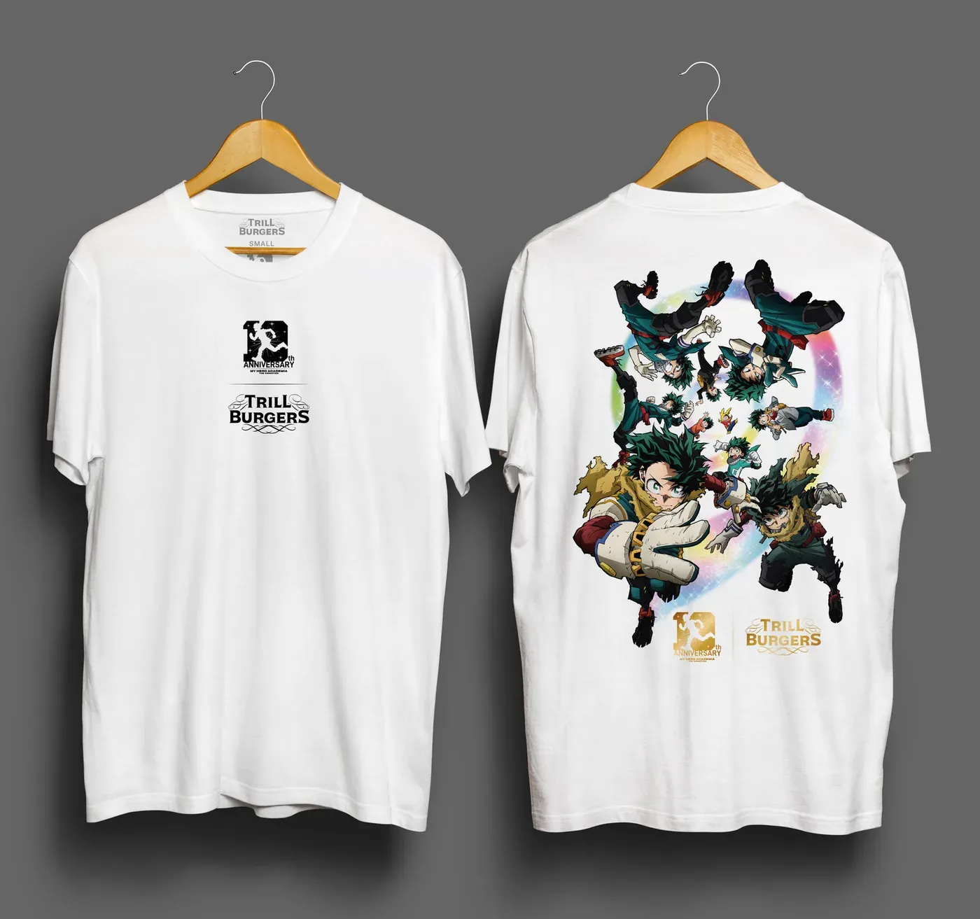 Two white t-shirts on wooden hangers against a grey background. The shirt on the left shows its front, with a small black 10th Anniversary My Hero Academia mark above a Trill Burgers logo on the chest, and a printed neck label reading Trill Burgers, Small. The shirt on the right shows its back, with a large full color print of green haired anime figures in tattered gold cloaks tumbling through a glowing rainbow ring, and a gold anniversary mark beside a gold Trill Burgers logo underneath.
